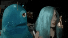 a woman with blue hair is standing next to a blue monster with a fish head .