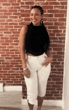 a woman standing in front of a brick wall wearing a black top and white pants