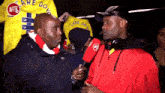 a man in a red jacket is talking to another man in front of a yellow flag that says " care do you "