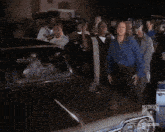a group of people are standing around a car and one of them is wearing a blue jacket