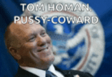 a man in a suit and tie is smiling in front of a flag that says tom homan pussy-coward