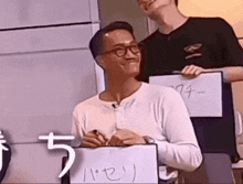 a man wearing glasses is writing on a piece of paper while another man holds a piece of paper .