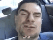 a man is sitting in the back seat of a car making a funny face and saying `` what '' .