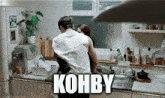 a man is carrying a woman in a kitchen and the word kohby is above them