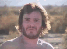 a man with long hair and a beard is standing in a field .