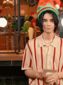 a young man wearing a striped shirt and a santa hat is standing in front of a christmas scene .