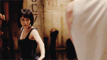 a woman in a black tank top stands in front of a wall with chinese characters on it