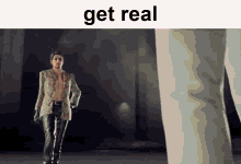 a man in a suit and leather pants is standing in front of a giant leg and says get real .