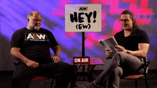 two men sitting in front of a sign that says hey