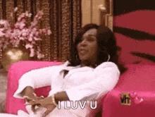 a woman is sitting on a pink couch and saying i love you .