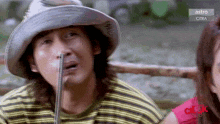 a man wearing a hat and a striped shirt has a snake sticking out of his nose while a woman looks on