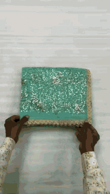 a person is holding a piece of blue fabric with sequins and a gold border .
