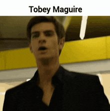 a man in a black suit is standing in front of a yellow wall with the name tobey maguire above him .
