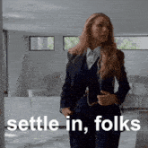 a woman in a suit is standing in a living room with the words " settle in folks " behind her