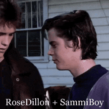 two young men are standing next to each other in front of a house and looking at each other .