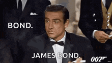 a man in a tuxedo and bow tie is sitting in front of a group of men in suits .