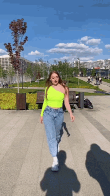 a woman wearing a neon green one shoulder top and blue jeans walks down a sidewalk