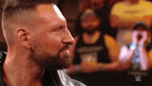 a man with a beard is wearing a black leather jacket with a w logo on his chest