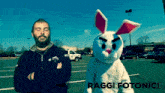 a man in a black jacket stands next to a stuffed bunny in a parking lot with the words raggi fotonici below him