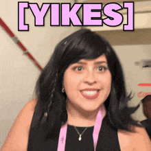 a woman wearing a black dress and a pink lanyard smiles in front of a sign that says [ yikes ]