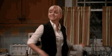 a woman in a vest and white shirt is dancing in the kitchen