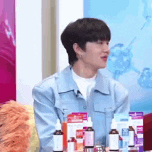 a young man in a blue shirt is sitting at a table with bottles of cosmetics .