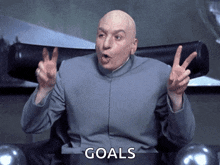 a bald man is sitting in a chair and giving a peace sign with the word goals behind him
