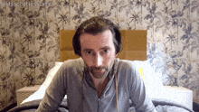 a man with a beard is sitting on a bed with a floral wallpaper behind him