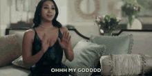 a woman in a black dress is sitting on a couch with her hands in the air and says ohh my godddd .