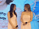 two women in white dresses are standing next to each other in front of a large screen holding microphones .