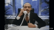 a man sitting at a desk talking on a phone with a gifrun.com logo on the bottom right