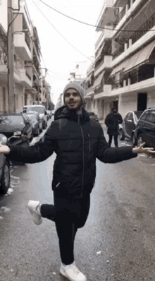 a man in a black jacket and a gray hat is standing on one leg on a city street .