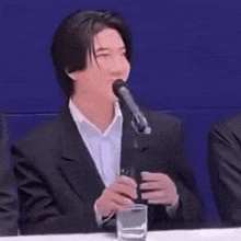 a young man in a suit is sitting at a table holding a microphone and a glass of water .