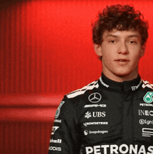 a young man wearing a petronas shirt stands in front of a red curtain
