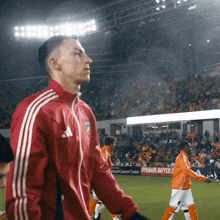 a man in a red adidas jacket stands on a soccer field in front of a sign that says dynamo cancer center
