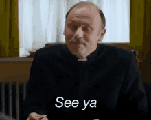 a man with a mustache is sitting at a table and says see ya .