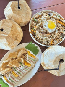 three plates of food are on a wooden table including a sandwich and noodles