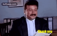 a man in a suit and tie is sitting at a desk holding a piece of paper and saying `` thank you '' .