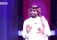 a man in a white shirt and a head scarf stands in front of a screen that says ' almajdchannels '