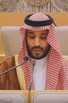 a man in a keffiyeh is speaking into a microphone in front of a sign with arabic writing on it
