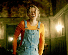 a woman wearing overalls and an orange shirt looks shocked