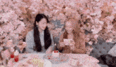 two women are sitting at a table with a cake and flowers on the wall behind them .
