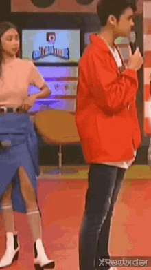 a man in a red jacket is talking into a microphone while a woman in a blue skirt stands behind him .