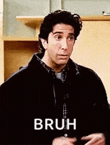 a man in a black jacket is standing in front of a wooden cabinet and says bruh .