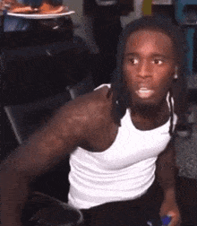 a man in a white tank top is sitting in a chair holding a remote control .