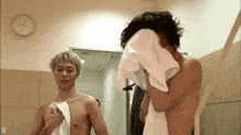 a shirtless man wipes his face with a white towel while another shirtless man looks on