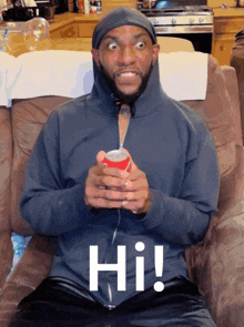 a man in a hoodie is sitting on a couch holding a can of soda and the word hi is on his shirt