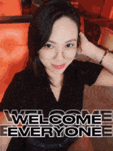 a woman wearing glasses is sitting in front of a sign that reads welcome everyone