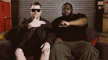 two men are sitting on a couch and one of them is pointing at the camera