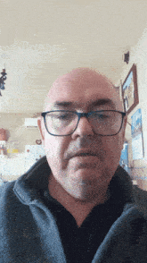 a bald man wearing glasses and a gray jacket looks at the camera